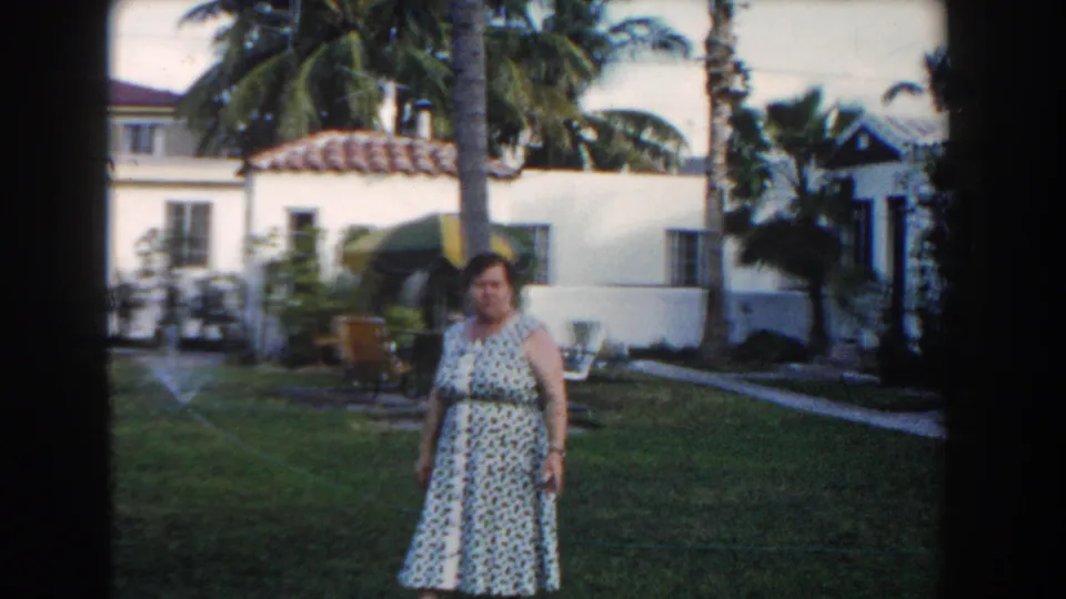 In 1960 Miami home-movie footage, an adult woman stands in a tropical residential garden with palms and a white stucco house behind her.