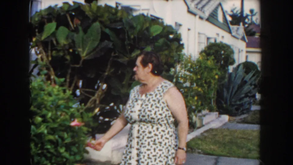 An adult woman in a patterned dress holds or touches a pink hibiscus near broad tropical leaves and a white residential building in Miami.