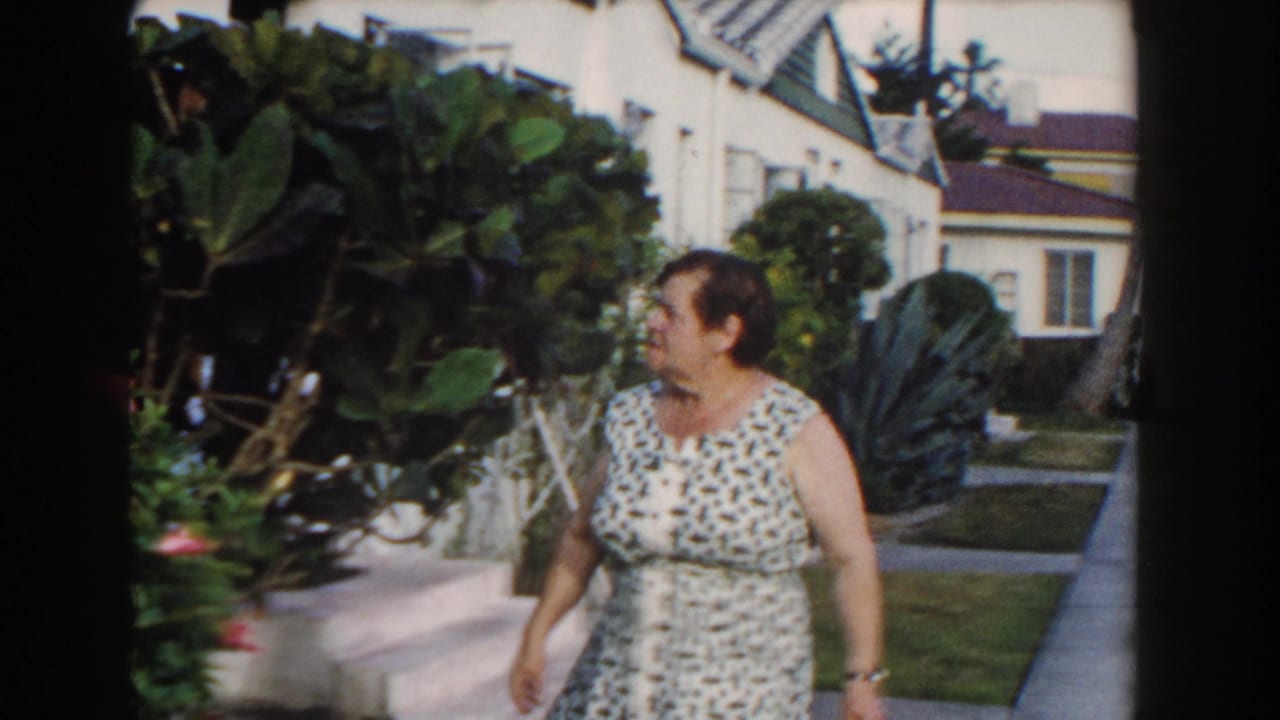 Woman walking past garden shrubs with white houses, lawn, and palms in the background.