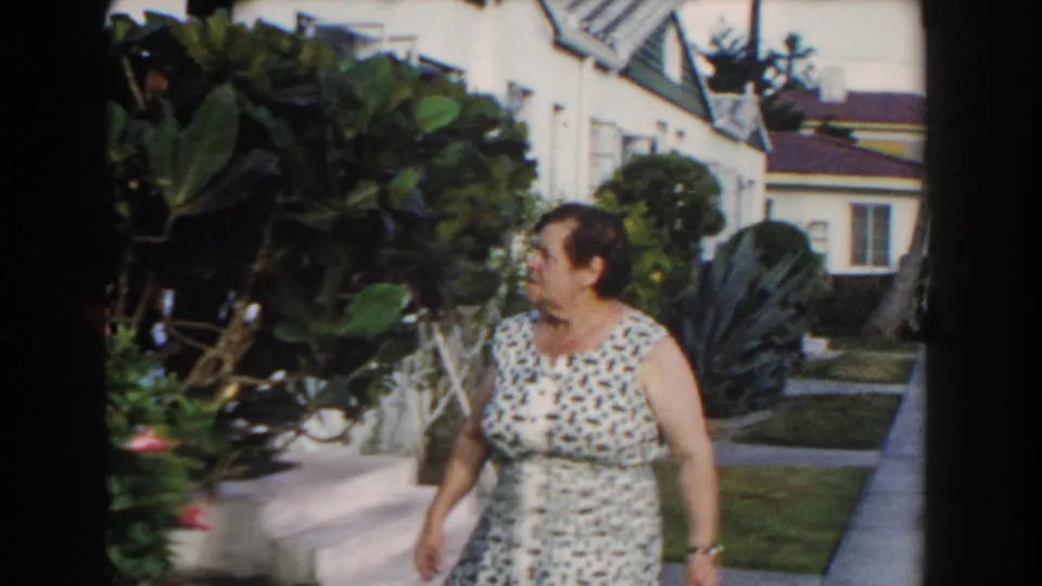 The woman walks along a Miami garden path with shrubs, lawn, palms, and white stucco houses behind her.