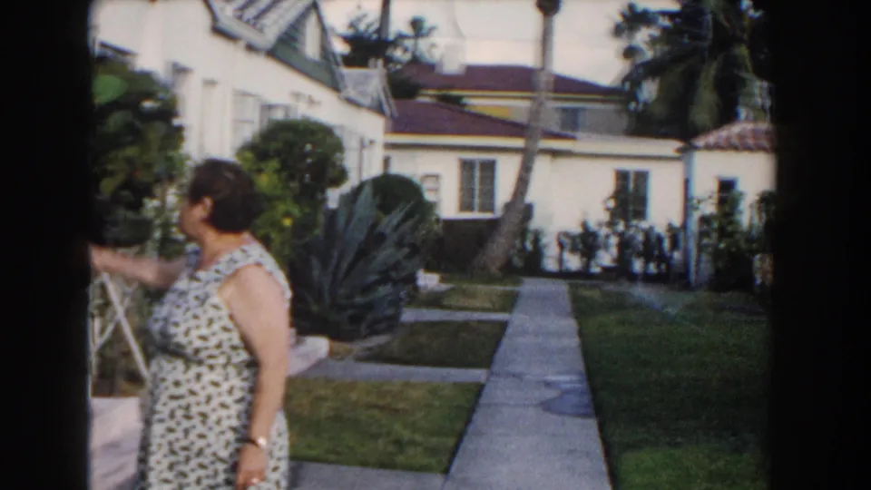 A side view shows the woman reaching toward foliage beside a concrete path in a 1960 Miami residential garden.