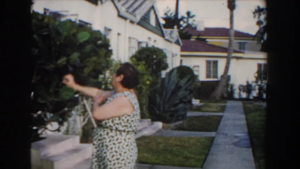 The woman reaches toward leafy garden branches beside a walkway, with Miami residential houses and palms visible behind her.