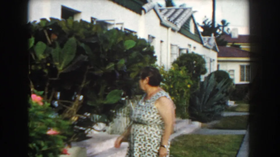 A woman in a patterned dress stands beside tropical plants in a Miami yard, with a white house and lawn visible behind her.