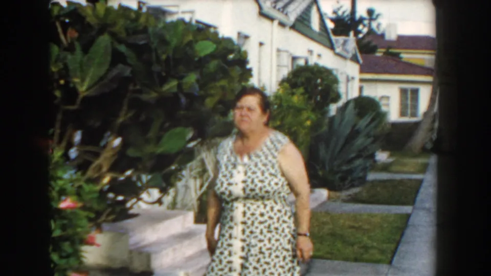 The woman walks near the garden path in a 1960 Miami residential yard, framed by tropical plants, lawn, and a white house.