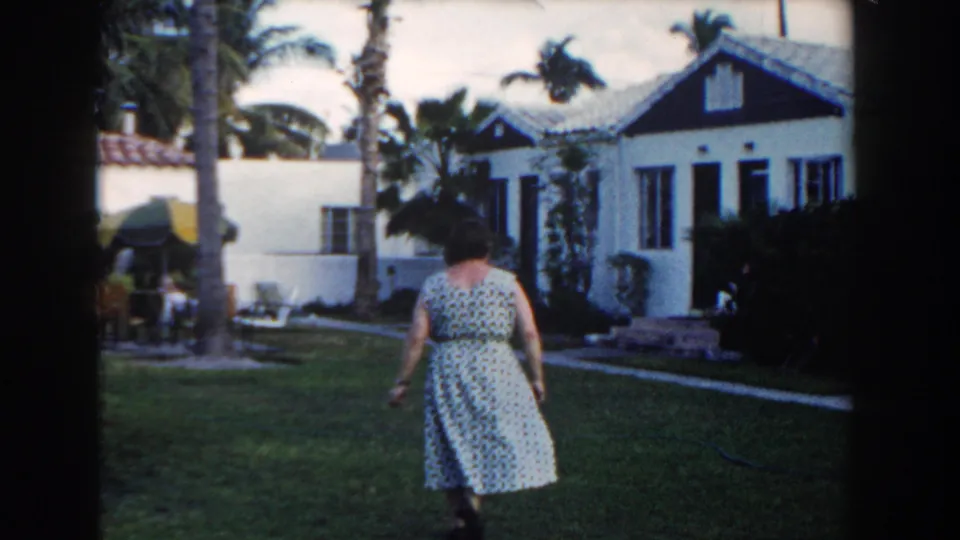 Seen from behind, the woman walks across a Miami lawn toward a white house, with palm trees and patio furniture in view.