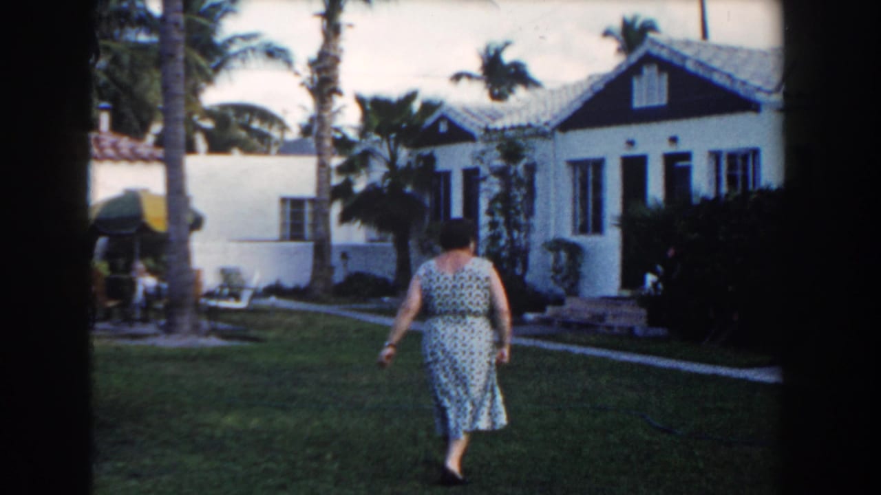 Woman in a patterned dress walking across a lawn toward a white bungalow.
