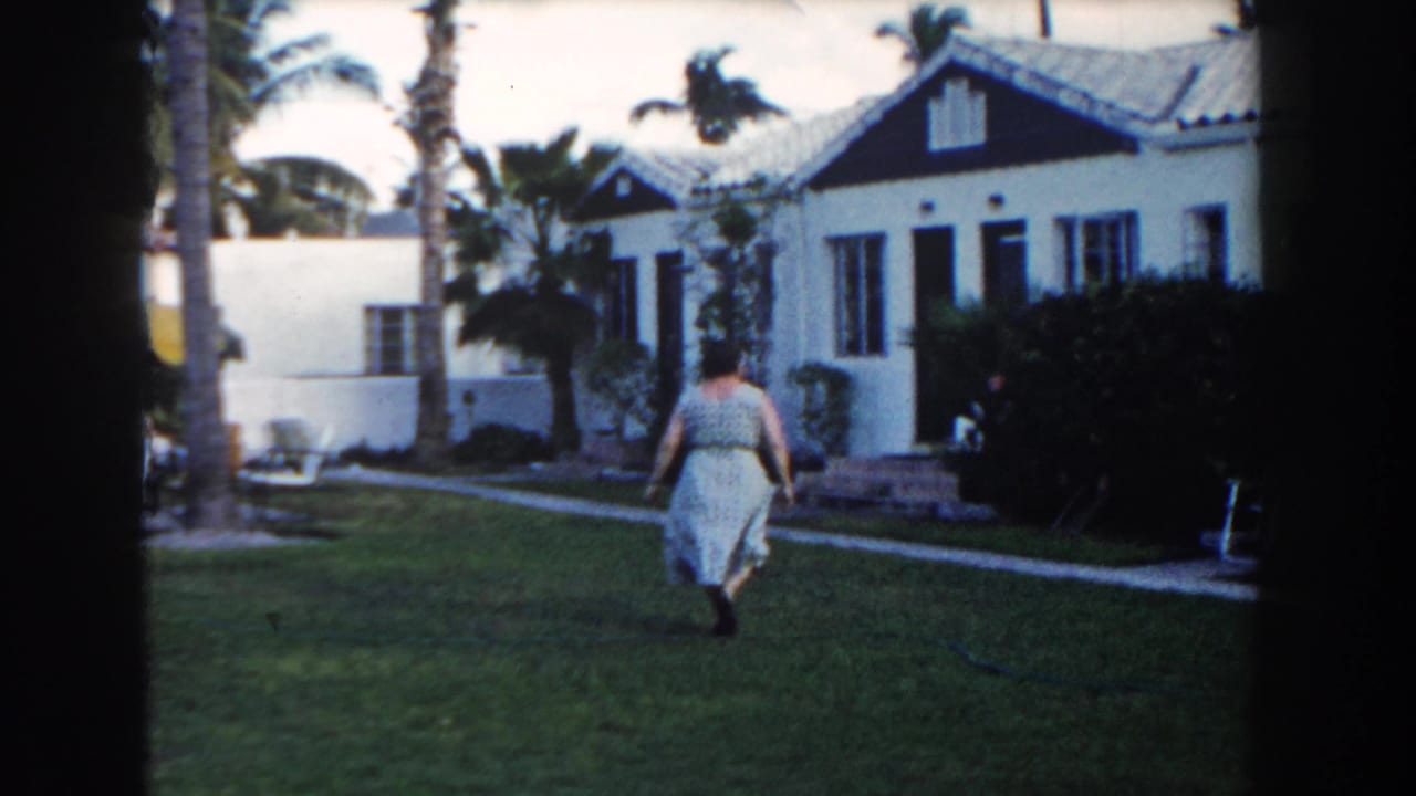 Woman walking farther across the lawn with a bungalow and palm trees ahead.