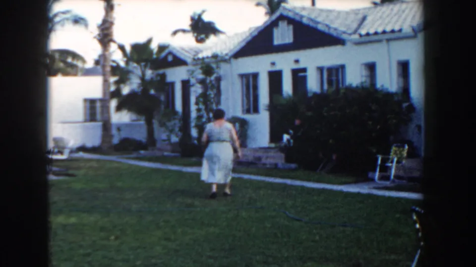 The woman approaches the bungalow entrance area, with shrubs, a walkway, palms, and a folding chair visible in the yard.
