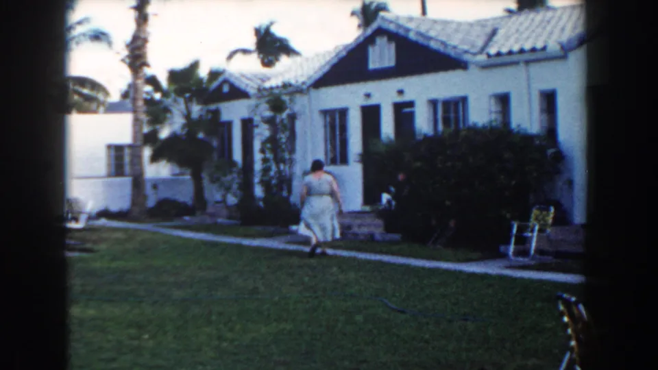 At the end of the clip, the woman is near the Miami bungalow exterior, with dark trim, palms, bushes, and a lawn chair in view.