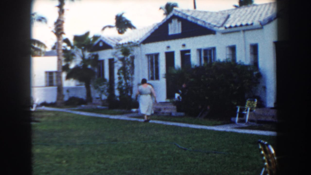 Woman in a light dress walks across a lawn path toward a white stucco house.