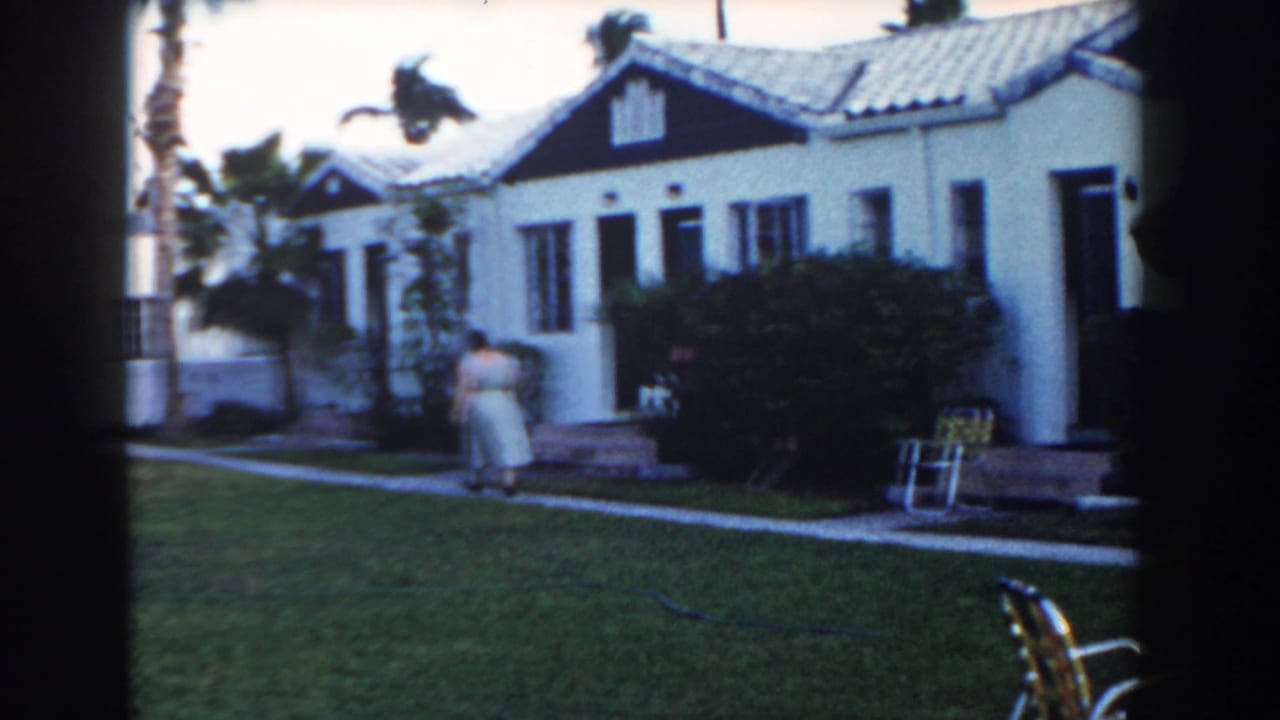 Woman stands near the front walk outside a white house with shrubs and a porch chair.