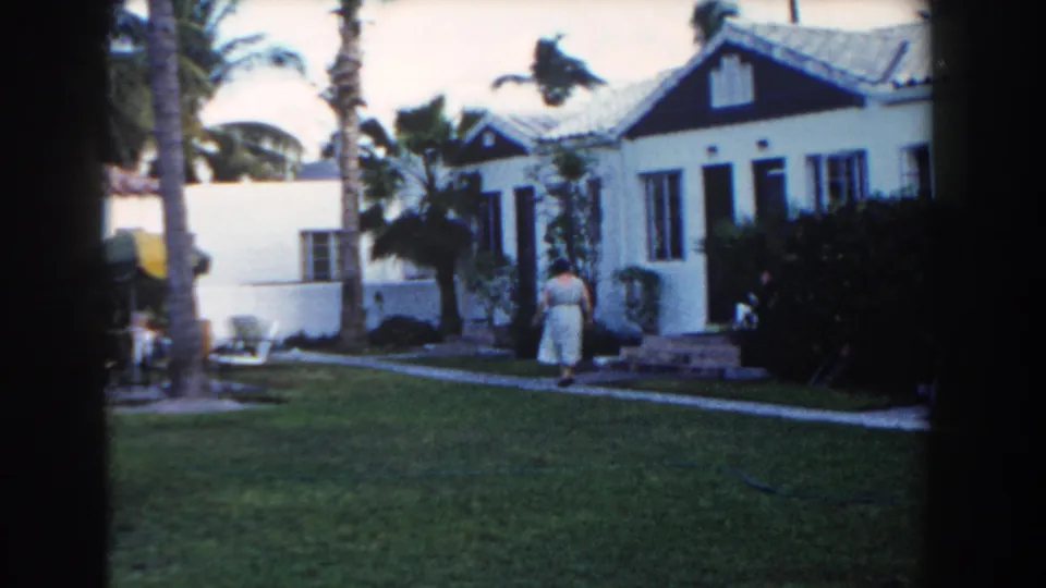1960 Miami home-movie still with a woman walking toward front steps at a white stucco house set behind lawn, palms, and shrubs.