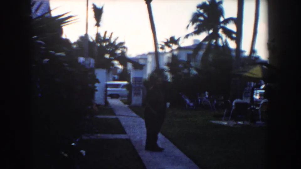 1960 Miami frame shifts to a darker garden view with a person by the walkway, palms overhead, lawn furniture, and a white vehicle beyond.