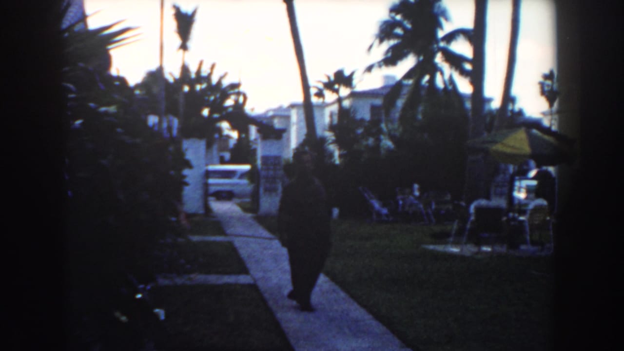 1960 Miami home-movie frame showing a man walking away along a residential sidewalk with palm trees, white buildings, lawn chairs, and a parked car.