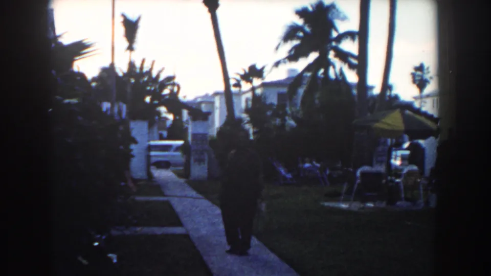 1960 Miami residential frame with a dark-clothed man walking away from the camera past palms, lawn seating, a yellow umbrella, and a parked car.