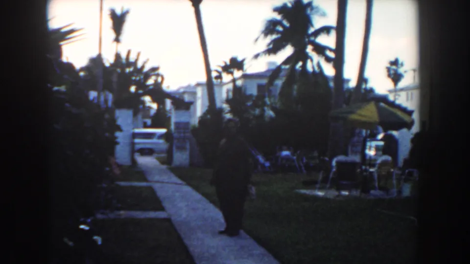 1960 Miami home-movie view of a man walking through a tropical residential setting with palms, white houses, lawn furniture, and a parked car.
