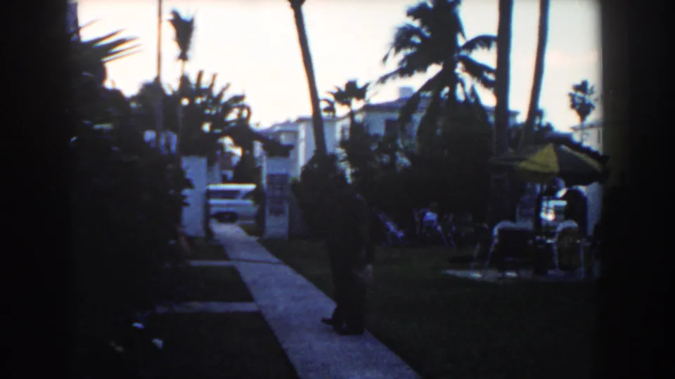 Final 1960 Miami frame showing the man on the sidewalk amid dense palms, residential buildings, lawn chairs, a yellow umbrella, and a parked car.