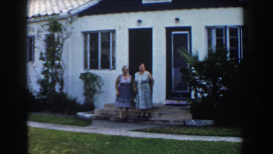 Two women in sleeveless dresses stand together on the steps of a Miami stucco house in 1960.