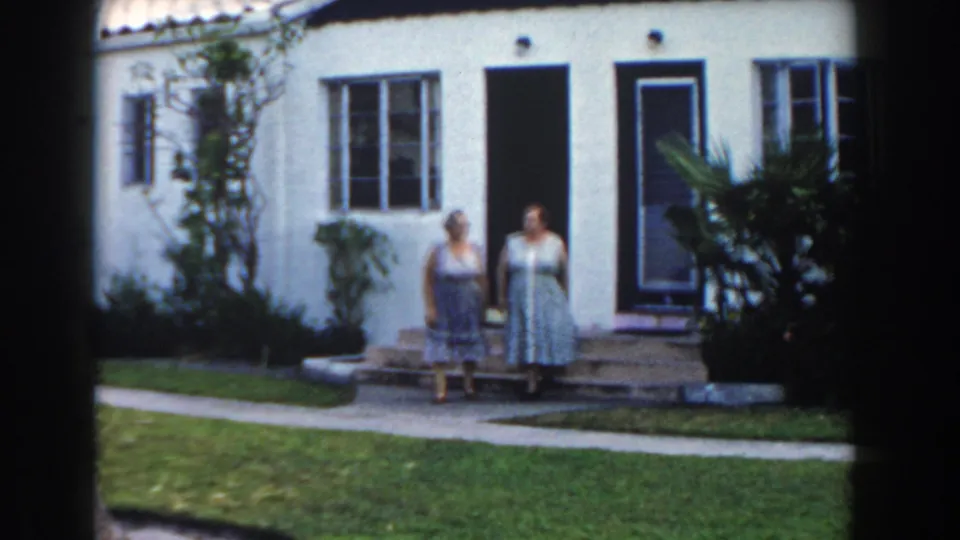 Two women stand outside a white stucco house with lawn and tropical plants in Miami, 1960.