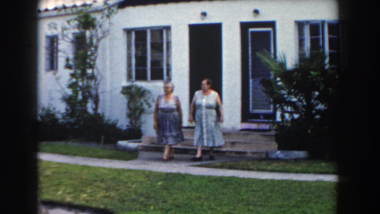 Two women stand near a white stucco house with a lawn and walkway.
