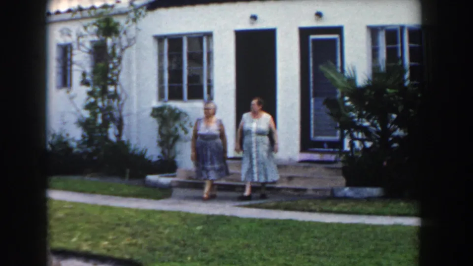 Two adult women in patterned dresses stand outside a Miami stucco house in 1960 home-movie footage.