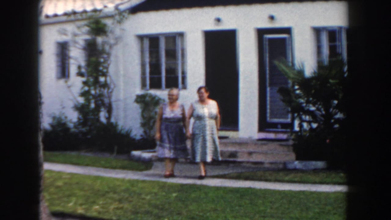 Two women walk together in front of a stucco house and yard.