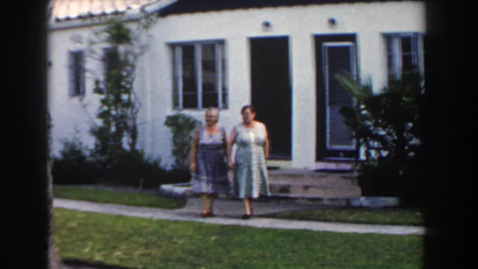 Two adult women walk hand-in-hand outside a Miami residential exterior in 1960 8mm footage.