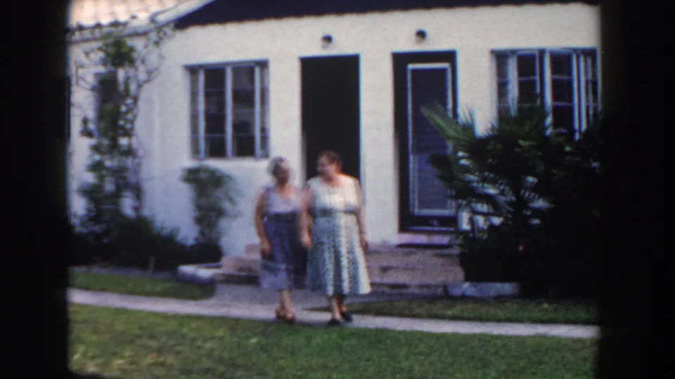 Two women in dresses move closer along a path in front of a Miami house, filmed on 8mm in 1960.