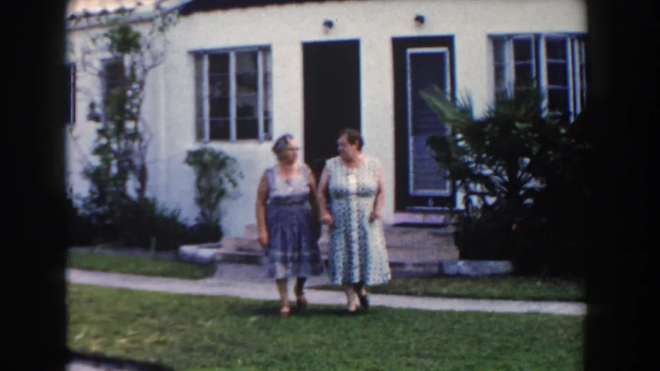 Two women continue hand-in-hand past the front yard of a Miami stucco house in 1960 home-movie footage.