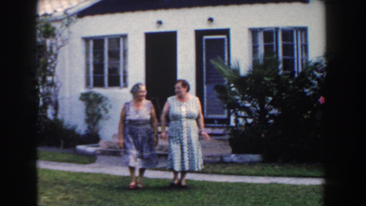 Two adult women in 1960s dresses stand together on a Miami lawn outside a white stucco residence.