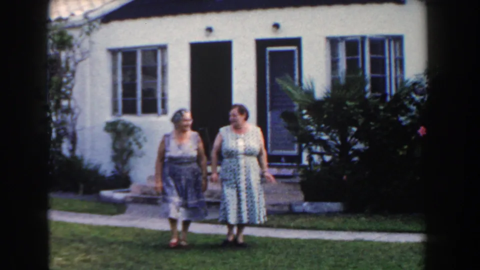 Two adult women in 1960s dresses stand together on a Miami lawn outside a white stucco residence.