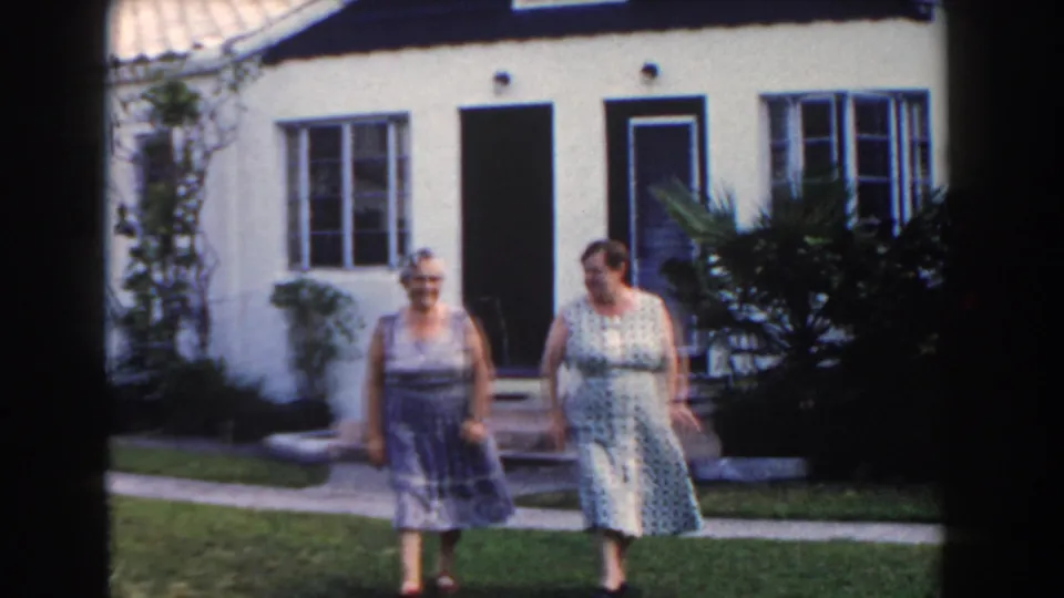 Two adult women in sleeveless dresses walk toward the camera outside a Miami stucco home in 1960.