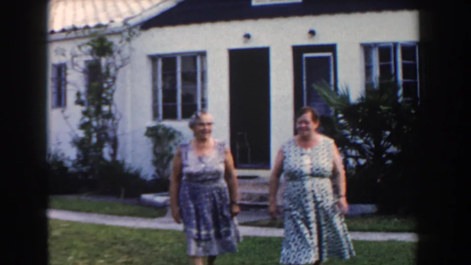 Two women in patterned dresses approach the camera on a residential Miami lawn with a stucco house behind them.