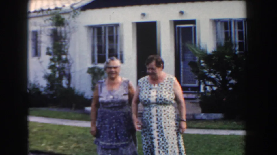 Two smiling adult women walk close to the camera on a Miami lawn, with a white stucco home and plants behind them.
