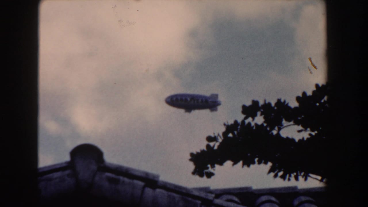 Goodyear blimp crossing cloudy sky above a tiled roof and tree branches.
