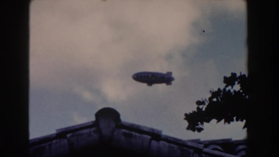1960 Miami frame showing the Goodyear blimp above a residential roofline and silhouetted foliage.