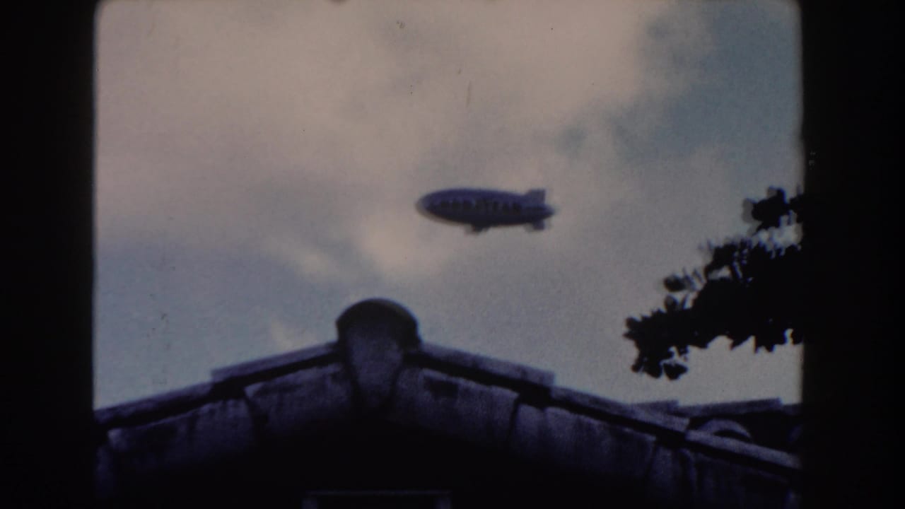 1960 Miami 8mm frame showing a Goodyear blimp passing above a silhouetted tiled roof and cloudy sky.