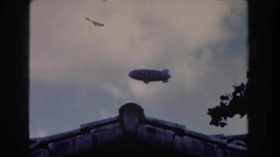 Goodyear blimp over a Miami roofline in a soft 1960 8mm home-movie frame, with tree branches at right.
