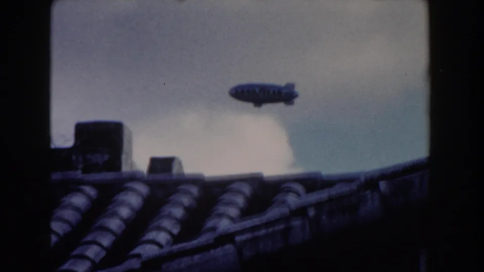 Goodyear blimp seen over a tiled Miami rooftop in a 1960 8mm frame, with chimney shapes and clouds below it.