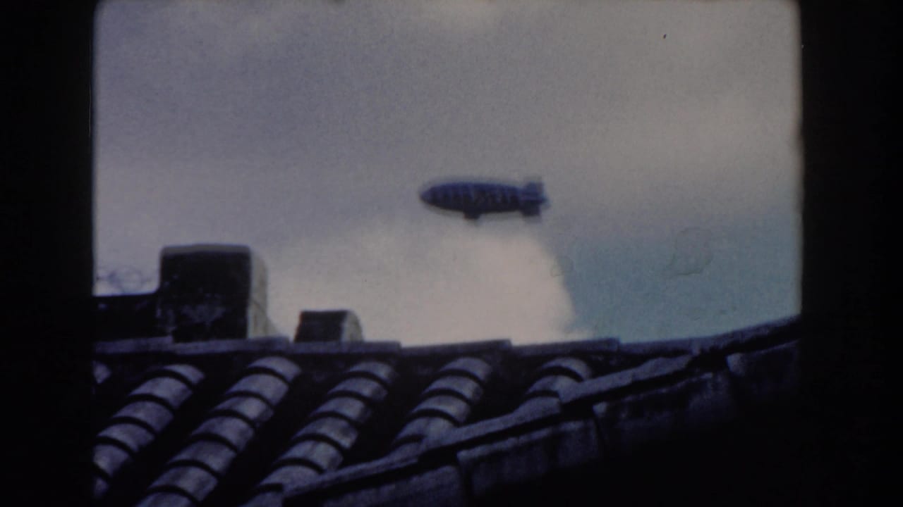 1960 Miami home-movie frame with a distant blimp passing over dark tiled rooftops beneath a cloudy sky.