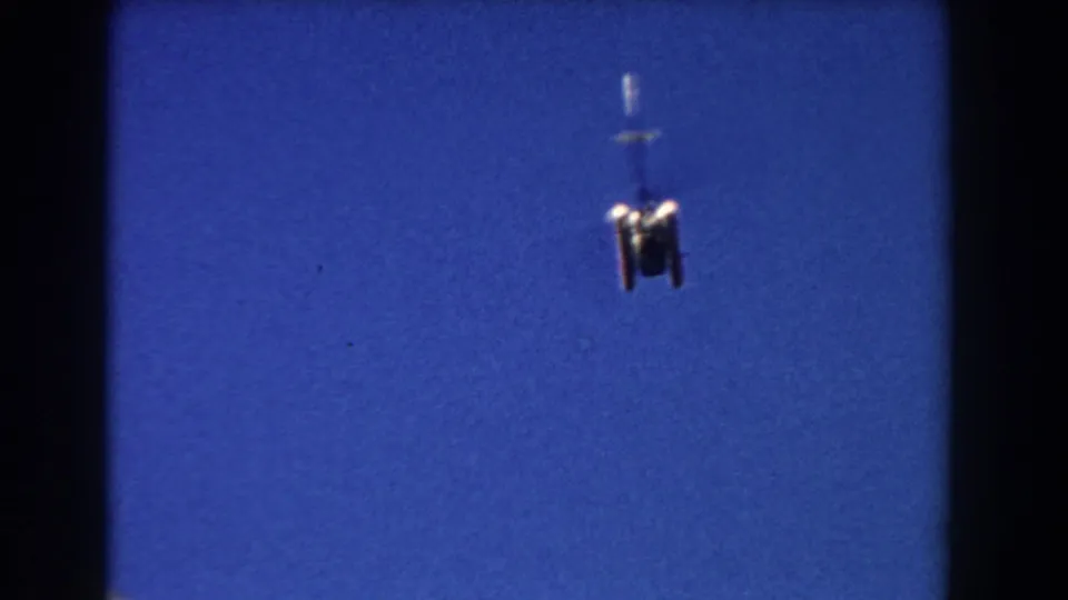1960 Miami home-movie frame with a small helicopter receding upward against a saturated blue sky.