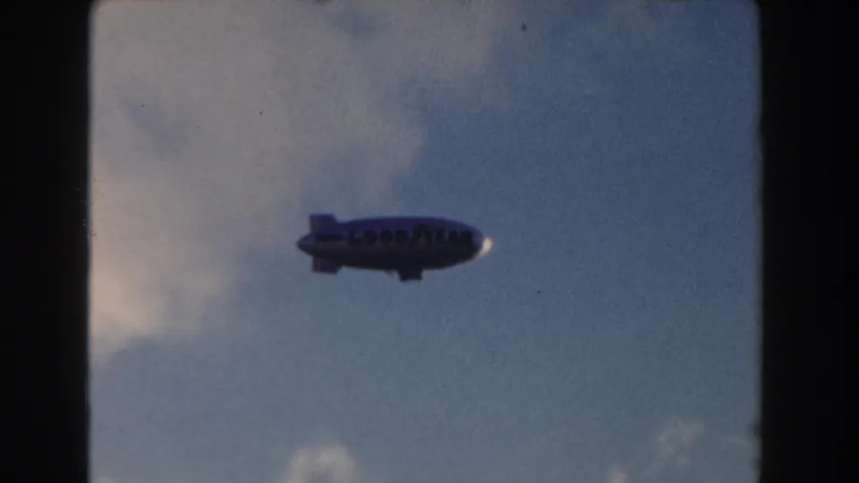 1960 Miami home-movie frame of a Goodyear-marked blimp crossing a blue sky with pale clouds.
