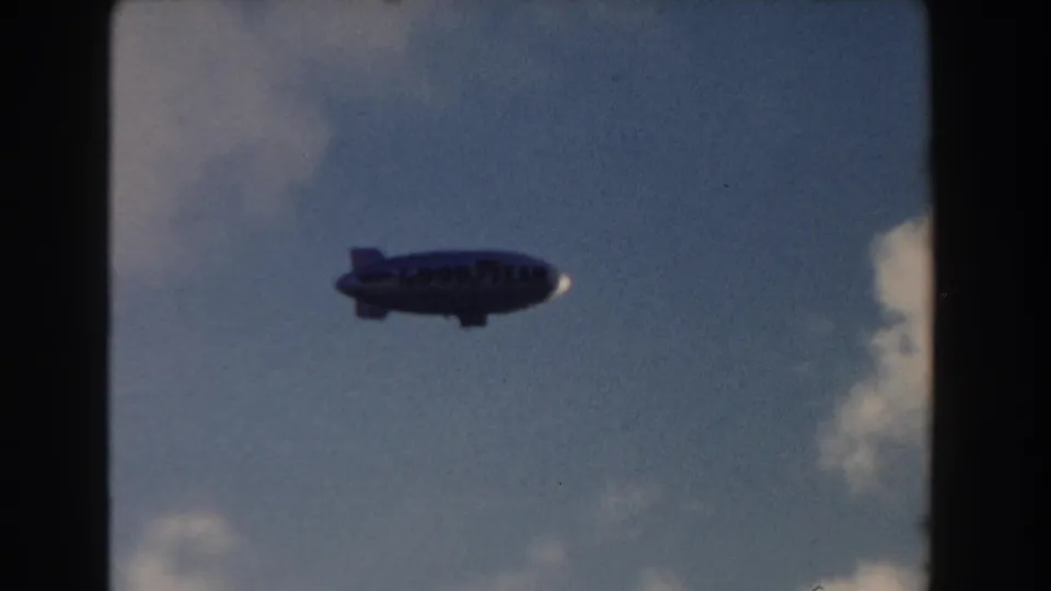 1960 Miami 8mm frame of a Goodyear-marked blimp in profile against a blue, lightly clouded sky.