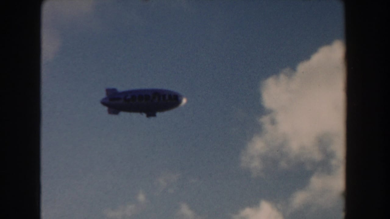 1960 Miami 8mm frame showing a Goodyear blimp moving across a partly cloudy blue sky.