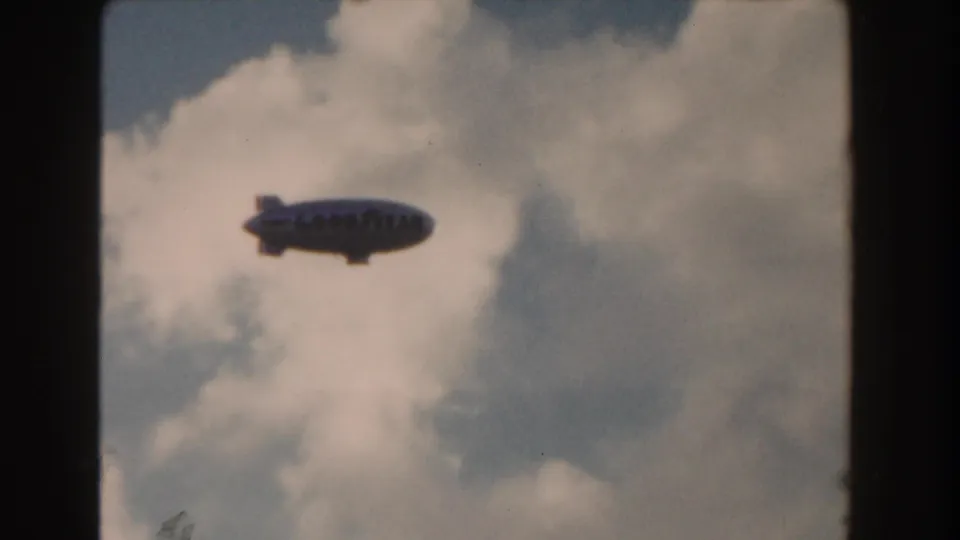 1960 Miami 8mm frame with a Goodyear blimp centered against pale blue sky and white clouds.