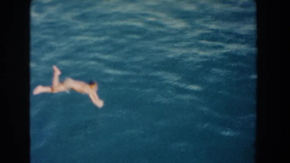 1960 Miami 8mm frame with blue water filling the view and a distant swimmer near the lower left, seen from an elevated or poolside angle.