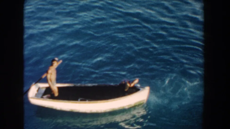 1960 Miami home-movie frame showing a man standing and waving in a small white boat as a swimmer reaches the side.