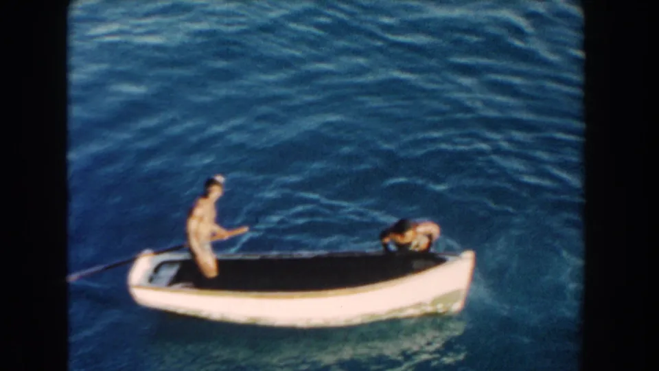 1960 Miami home-movie frame of two people in a small white boat on rippled blue water, with one person standing near a paddle.