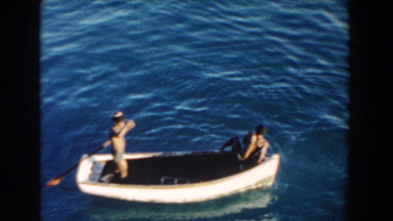 A person stands with a paddle while another sits in a small white boat on blue water.