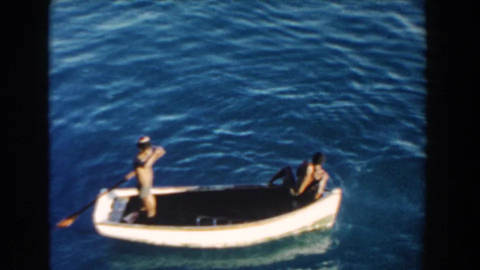 1960 Miami frame showing a person standing with a paddle and another seated at the stern of a small white boat on open blue water.